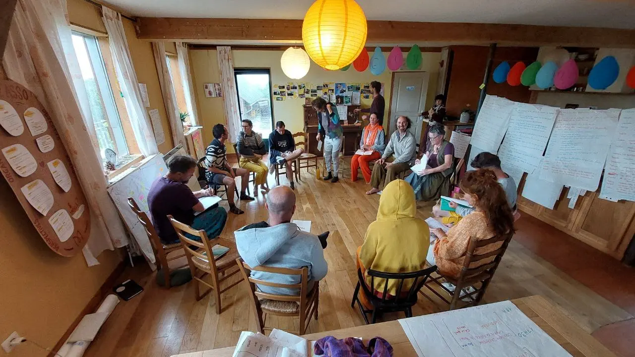 Une quinzaine de personnes en cercle dans une salle, carnets sur les genoux, affiches au mur